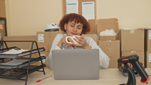 Woman holding coffee mug with both hands at laptop desk in building surrounded by cardboard boxes and parcel delivery; contentment.