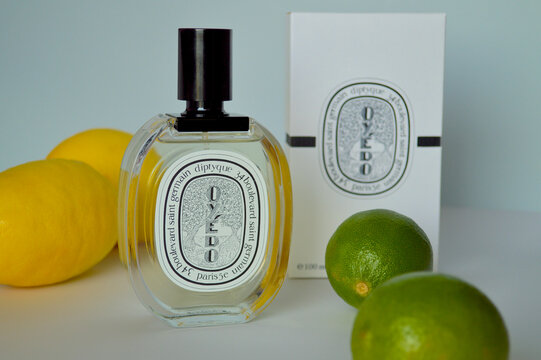 Luxury Diptyque Oyedo perfume bottle and white packaging box arranged with fresh yellow lemons and green limes on a clean studio surface for a citrus fragrance concept.