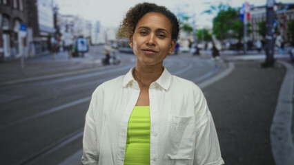 Fototapeta premium Woman with visible face and bare neck smiling, standing midblock on tram tracks on an urban street wearing a white shirt and neon green top; confidence resilience strength.