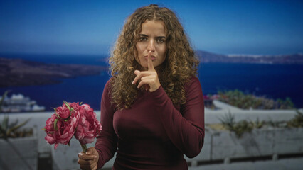 Obraz premium Woman with curly hair holds pink flowers and presses index finger to lips on a building terrace overlooking the sea; silence secrecy.