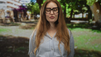 Woman wearing black glasses and a striped blouse stands with eyes closed among green trees in park  calm reflection. © Krakenimages.com