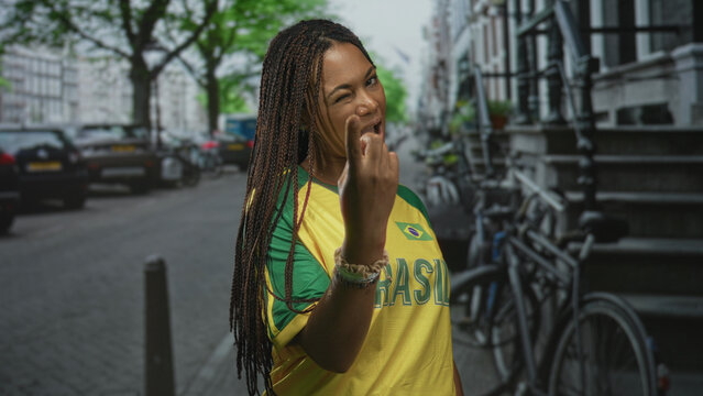Woman beckons with finger on city street wearing yellow brazil jersey with flag visible; confidence invitation.