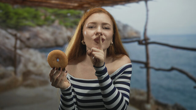 Woman redhead holding doughnut, finger to lips and bare shoulder visible in studio; playful indulgence.