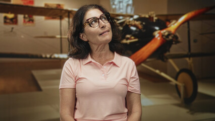 Middle age woman with glasses tilts head and looks up at vintage airplane propeller in museum building  quiet curiosity. © Krakenimages.com