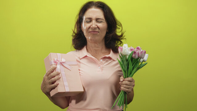 Woman holding pink gift box and bouquet with hands, eyes closed and slight grimace in studio; bittersweet memory reflection.