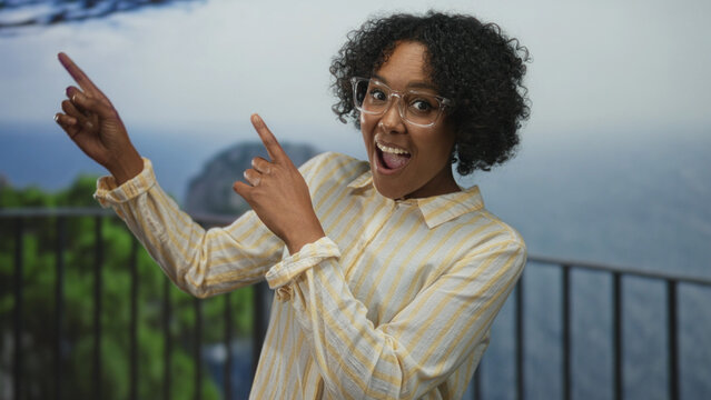 Young african american woman in yellow shirt and glasses pointing finger to ocean view on street balcony; excitement.