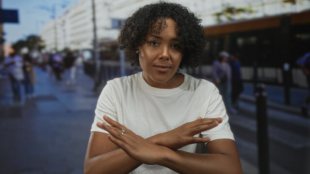Young african american woman in white t shirt crossing arms in front of blurred urban buildings on street; denial.