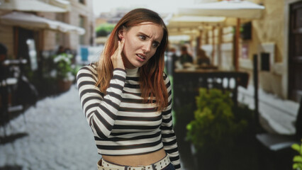 Fototapeta premium Woman holding hand to neck touching with a striped crop top exposing midriff on a cobbled street outside a cafe with umbrellas and tables; discomfort.