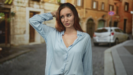 Fototapeta premium Woman showing slight cleavage shrugging both hands on a narrow cobblestone street with parked car and old building; casual bemusement.