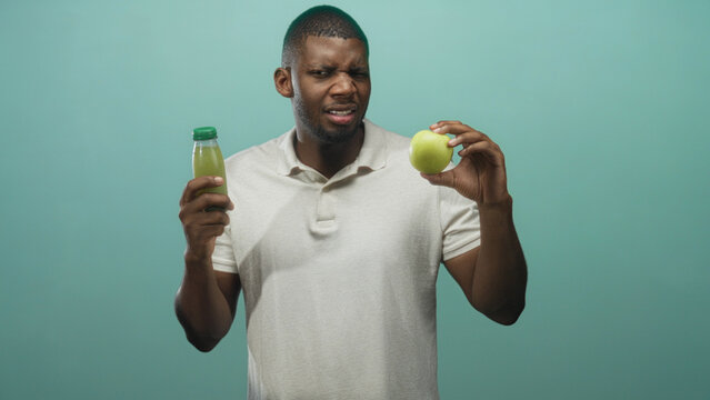Man in beige polo grimaces with closed eyes while holding green apple and bottled juice in a mint studio; taste test skepticism.
