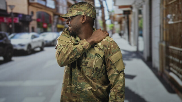Man soldier in camouflage uniform and cap rubbing shoulder, wincing from backache while standing on a city street near parked cars; duty fatigue resilience.