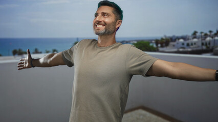 Obraz premium Man with arms outstretched showing hands on building rooftop terrace overlooking sea, smiling and looking up; joy escape calm.