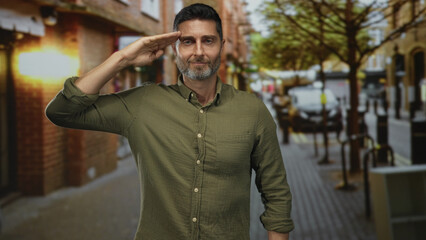 Fototapeta premium Man salutes with hand to forehead on a city street with slight smile while wearing green shirt; quiet respect.