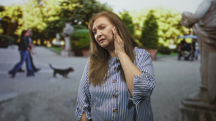 Fototapeta premium Senior woman clutching her ear, eyes closed and wincing on street with statue and trees; aging discomfort.