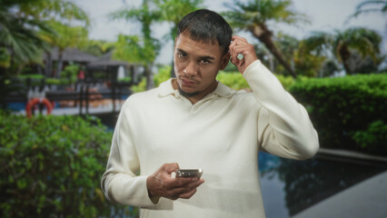 Fototapeta premium Man holding smartphone and scratching ear by a resort pool with palm trees, lifebuoy and bungalow, wearing a cream sweater and looking at phone; confusion.