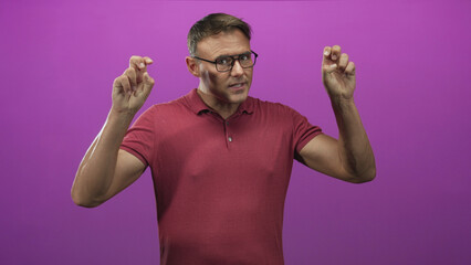 Fototapeta premium Man wearing glasses and red polo showing crossed fingers with both hands, hands raised in a purple studio setting; hopeful luck.