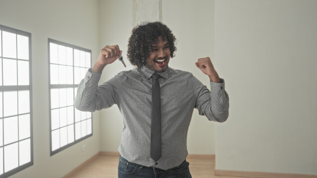 Man holding house keys in hand, smiling and raising fist beside tall windows in empty building room with wooden floor; homeownership pride.