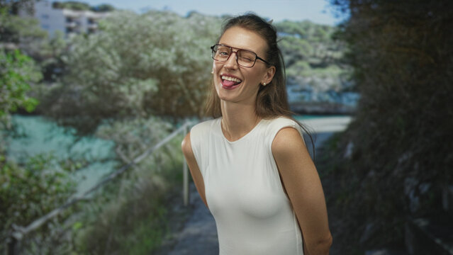 Woman with bare shoulders wearing glasses smiling and sticking tongue out on a shaded forest path by green water; playful joy.