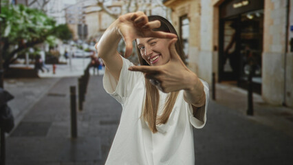 Obraz premium Woman in white t shirt framing her face with her hands, winking and smiling on a cobblestone street; creative joy.