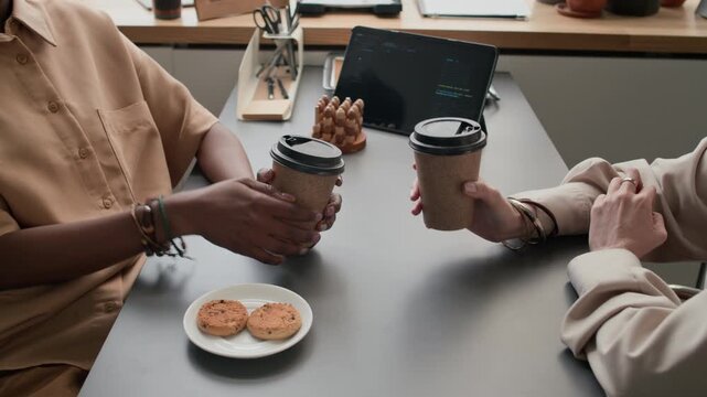 High angle close up shot of two unrecognizable women enjoying coffee and cookies during lunch break at office table