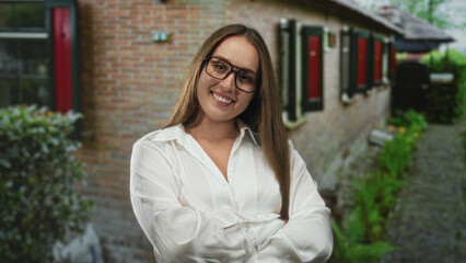 Obraz premium Woman wearing glasses smiling with arms crossed outside a brick building entrance; confidence welcome home.