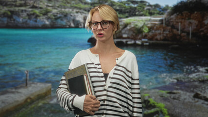 Fototapeta premium Woman holding book and notebooks, touching neck and adjusting glasses, striped sweater, posed before seascape backdrop in studio; thoughtful reflection.