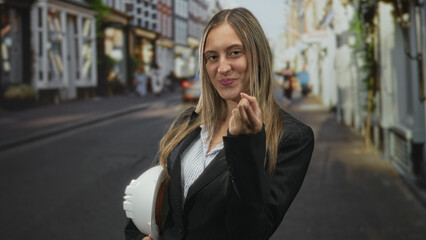 Fototapeta premium Young woman in blazer holding white hardhat under her arm and smiling toward camera on a narrow european street; confidence career ambition.