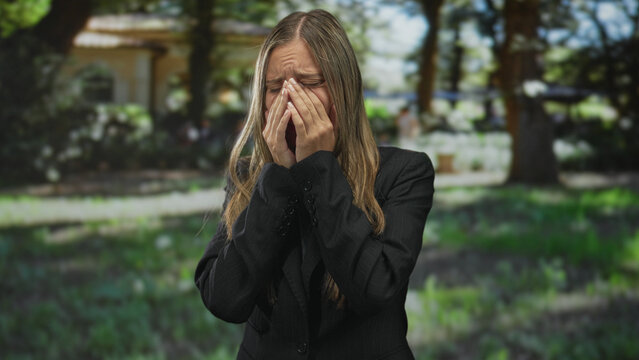 Woman covering face with hands and rubbing her eyes while crying in front of a building and trees in an outdoor courtyard; grief loss.