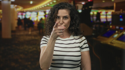 Fototapeta premium Young hispanic woman stares at camera and points finger at slot machine in casino building; determination.