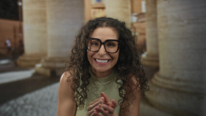 Fototapeta premium Woman wearing eyeglasses in a sleeveless top clapping hands on a cobbled street near a historic building; celebration joy.