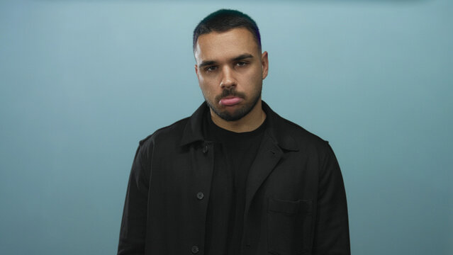 Hispanic man with beard looks down revealing face and lips in blue studio; melancholy reflection doubt.
