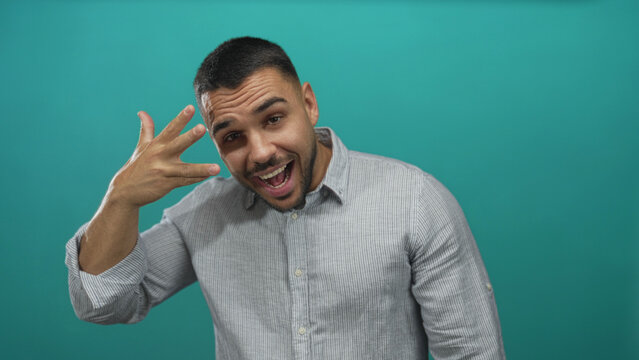 Young hispanic man covers eyes with hand and smiles broadly against a bright teal studio wall; playful shyness.