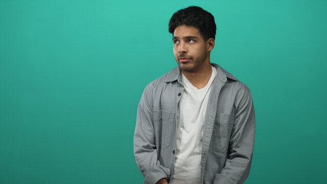 Young hispanic man in casual shirt with hand on chest and hesitant expression in teal studio setting; apologetic regret.