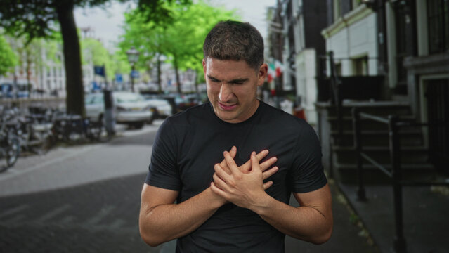 Young hispanic man grimacing clutching chest with bare hands on city street near bicycles and parked cars; heartattack emergency pain.