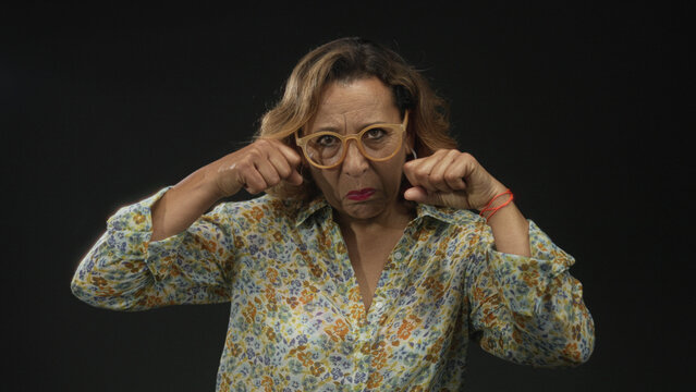 Middle age woman holding fists near face while frowning and wearing eyeglasses and floral shirt in black studio; frustration.