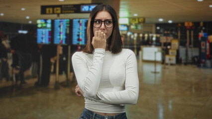 Fototapeta premium Young woman wearing glasses biting her nails with hand to mouth gesture in an airport terminal near departure screens and queues; travel anxiety delay.