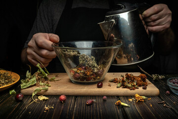 A person pours hot water from a kettle into a bowl filled with dry herbs and flowers. Various dried plants are scattered on the table around them. The work takes place in a dark kitchen © Віталій Б.