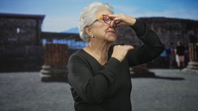 Elderly woman pinches her nose and adjusts eyeglasses while wearing a black sweater in a building among ruins with a distant crowd; disgust.
