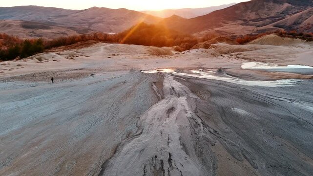 Aerial view of the Vulcanii Noroiosi Paclele Mici mud volcanoes with grey mud flows, rolling hills and a lone person at sunset in Scortoasa, Buzau County, Romania.