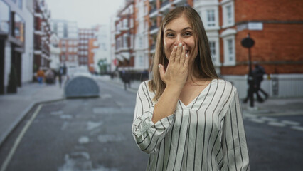 Fototapeta premium Young blonde woman covering mouth with hand on bustling city street beside brick building facade under daytime light; surprise.