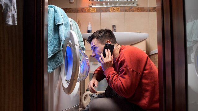 Man kneeling in a small bathroom, looking inside the washing machine with its door open while making a phone call, seeking assistance for a broken appliance