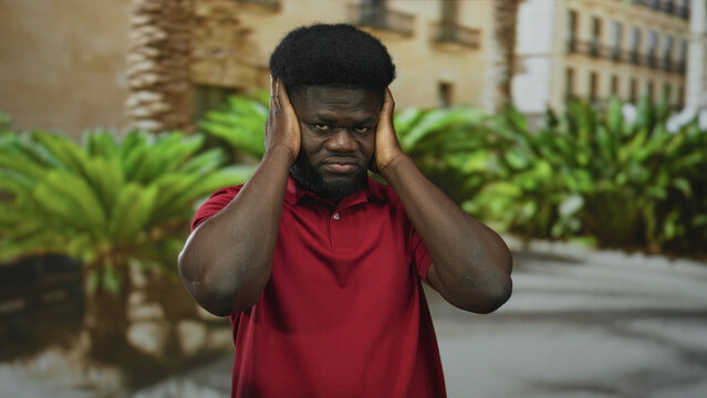 Man covering ears with hands in front of building courtyard wearing red polo shirt and frowning in sunlight; sensory overload frustration.