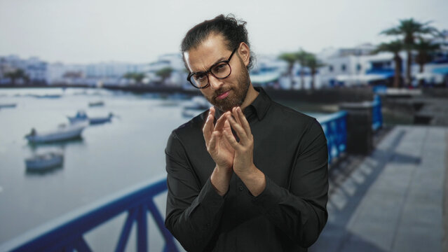 Man claps hands on seaside promenade street while peering at palms and wearing glasses and black shirt; quiet contemplation.