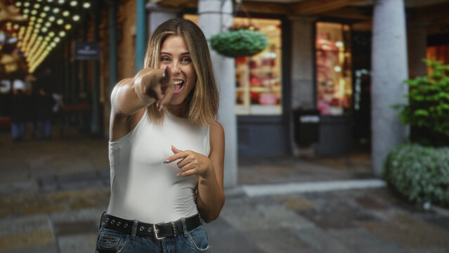 Woman points finger at camera on a street storefront with string lights, wearing white sleeveless top and jeans; playful.