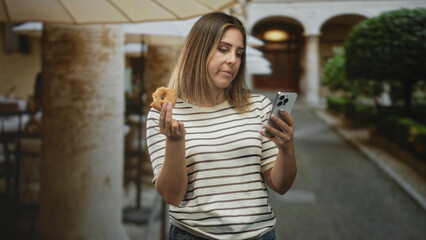 Fototapeta premium Woman holding a donut in her hand and checking a smartphone while standing under a colonnade by a street; distraction.