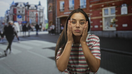 Fototapeta premium Young woman holds hair between fingers and inspects roots while standing on a busy city street crosswalk by brick buildings wearing a striped white and red shirt; frustration.