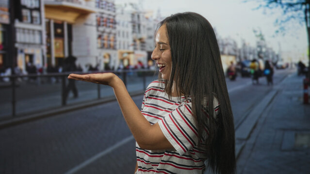 Young hispanic woman presenting palm up in striped shirt on busy city street while smiling and looking at hand; joy playfulness.