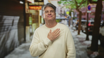 Fototapeta premium Man hand to chest on street in urban storefront scene wearing cream sweater, looking thankful with slight smile; gratitude reflection.