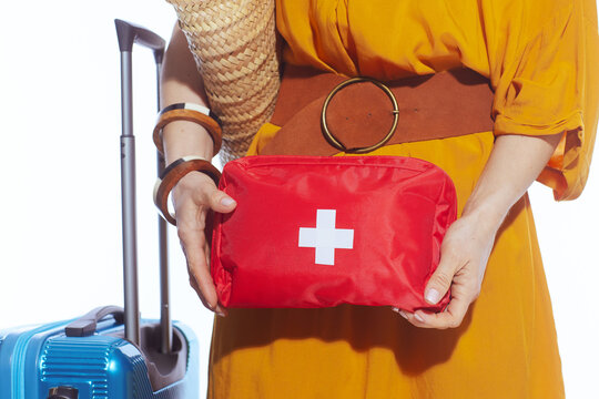A woman in a vibrant yellow dress prepares for her summer vacation by packing a red medical first aid kit next to her blue hardshell suitcase, prioritizing travel health and safety.