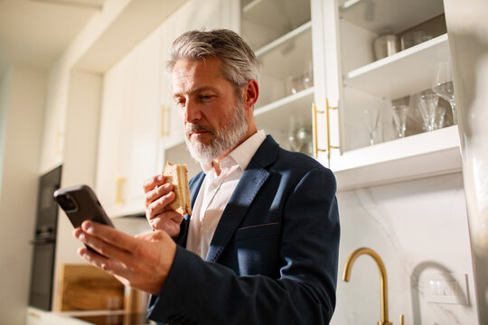 Mature businessman eating sandwich and using smartphone in modern home kitchen